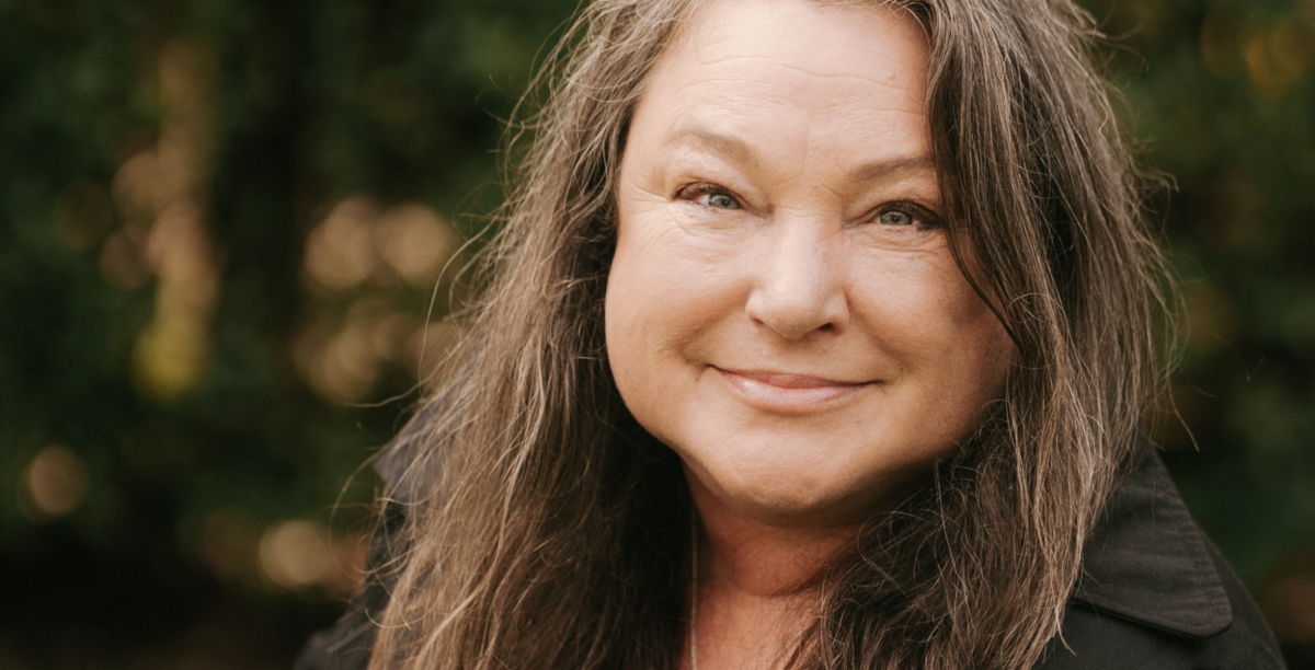 Julie is outside in front of a wall of greenery. She smiles over her left shoulder towards the camera in a close head shot. 