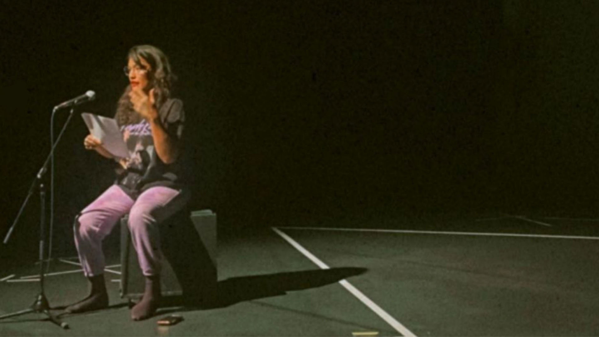 Pelenekeke is seated on stage, with a long shadow behind her.