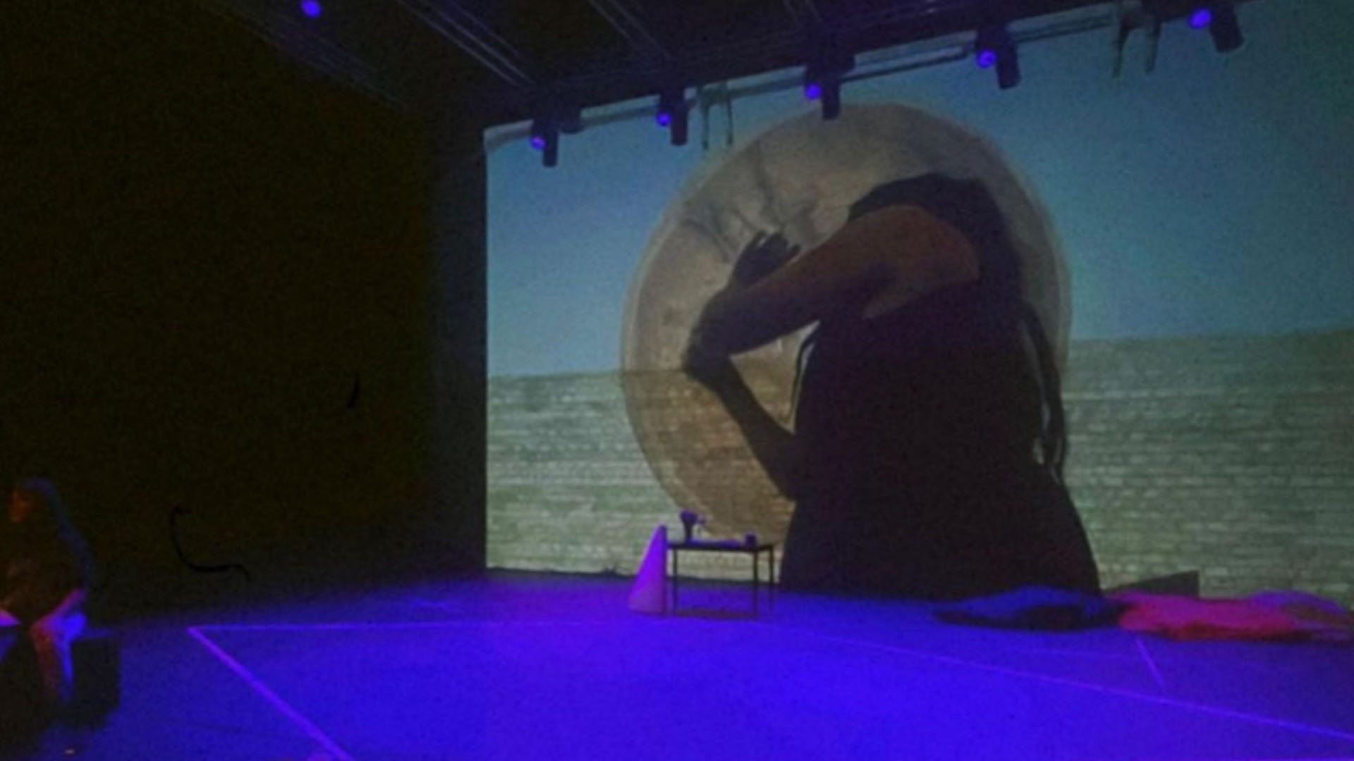 A dark theatre stage with a brick wall. The floor is blue and there is a projection of a woman's body on the wall.