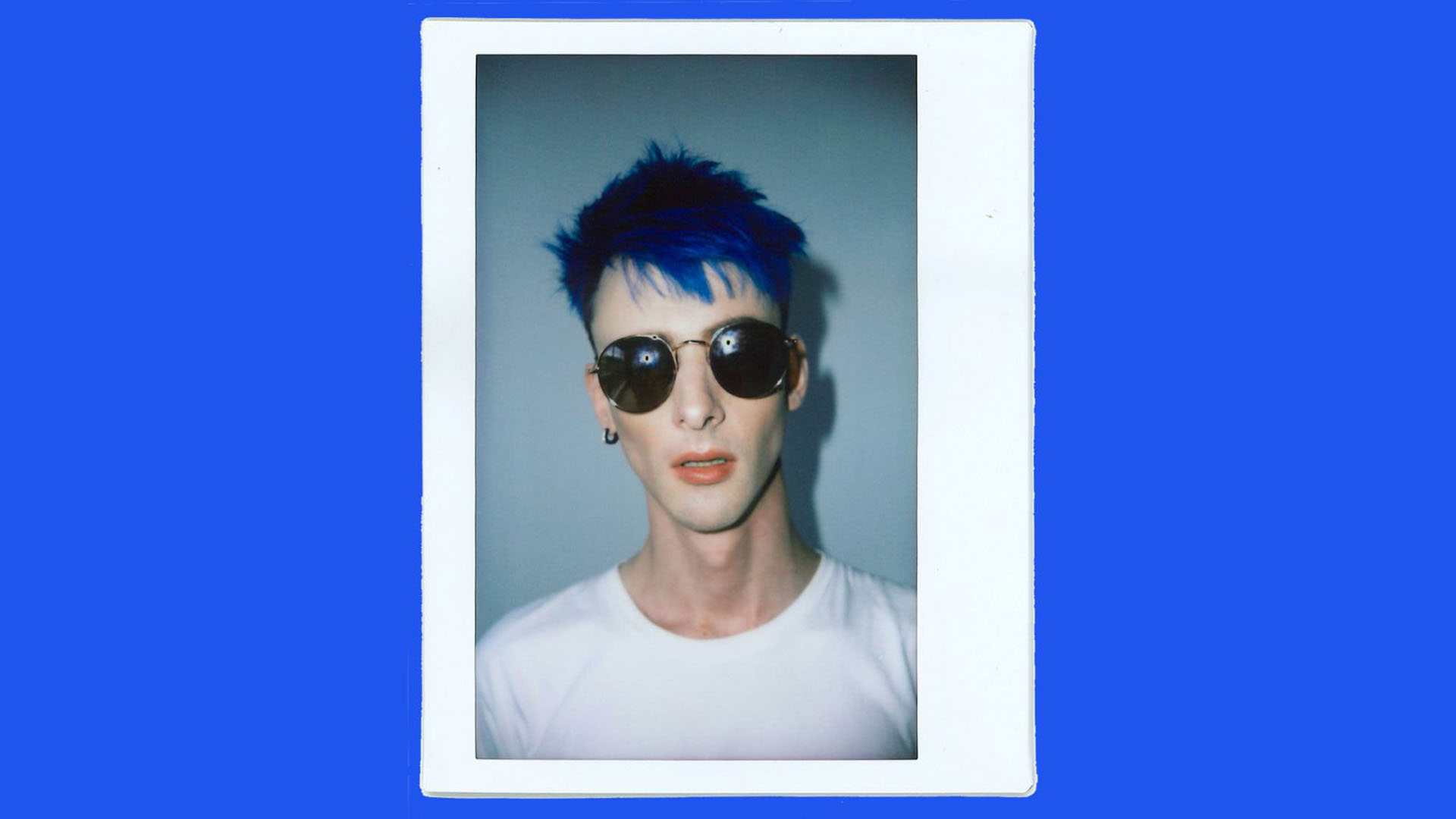 A close up photo of Ari's face, who has blue hear and is wearing round sunglasses. 