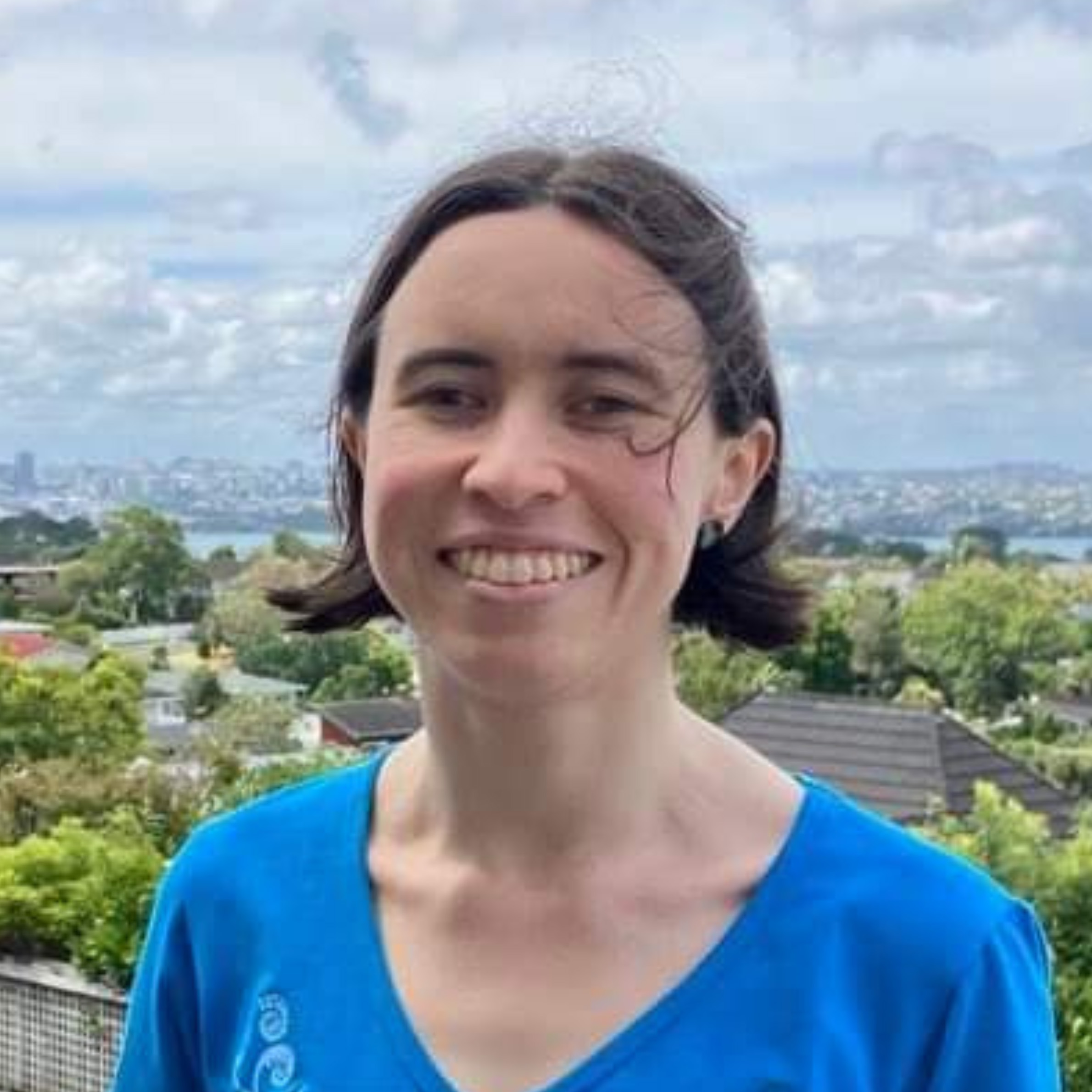 Áine stands smiling outdoors on a house balcony, their short brown hair blowing in the summer breeze. They are a white non-binary femme. Suburban treetops dominate the background, with Auckland's skyline visible in the distance.