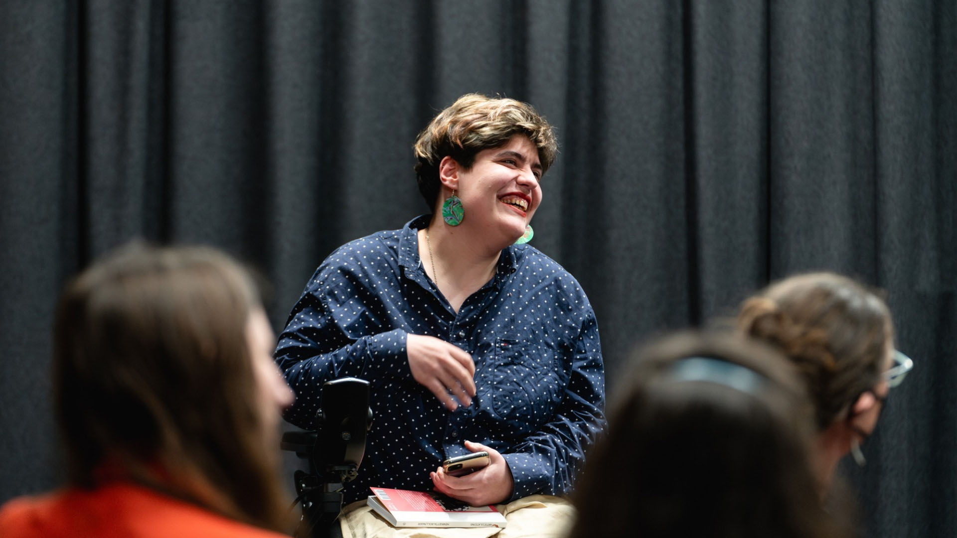 Henrietta is a pākehā person with short brown hair with highlights and sitting in a powered wheelchair. They are smiling away from the camera in front of an audience.