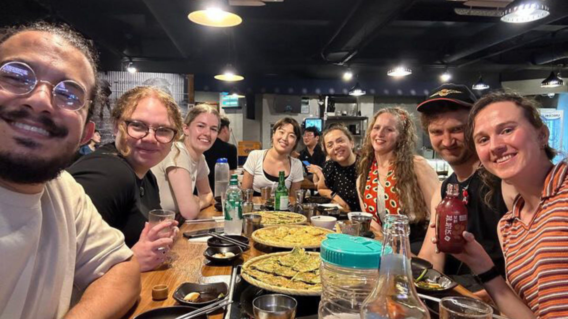 Eight young people are sitting at a restaurant table enjoying a meal and drinks.