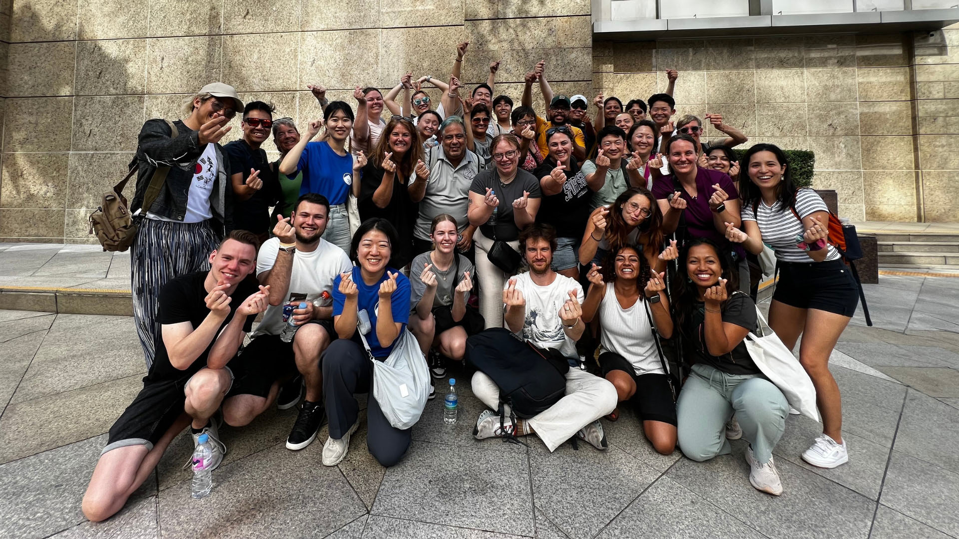 A group of around 30 young people smile at the camera and make a heart shape with their hands