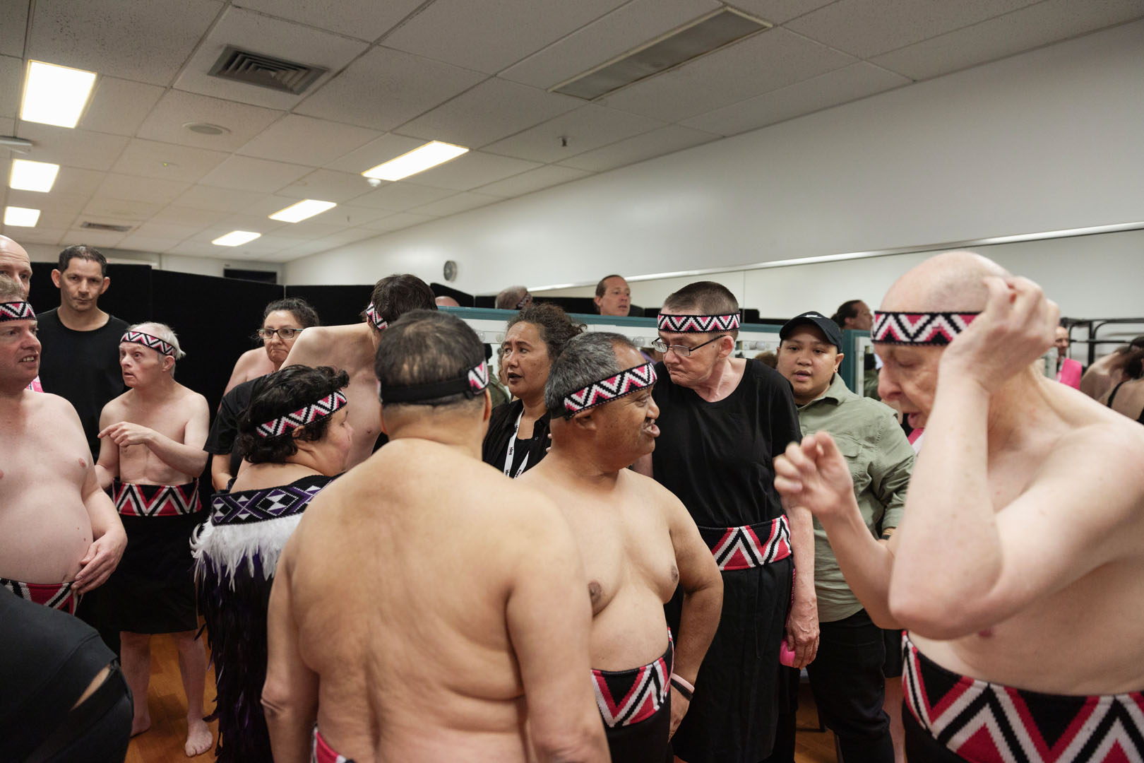 A group of kaihaka are backstage getting ready to go on stage, they are wearing tīpare and some male performers are shirtless. 