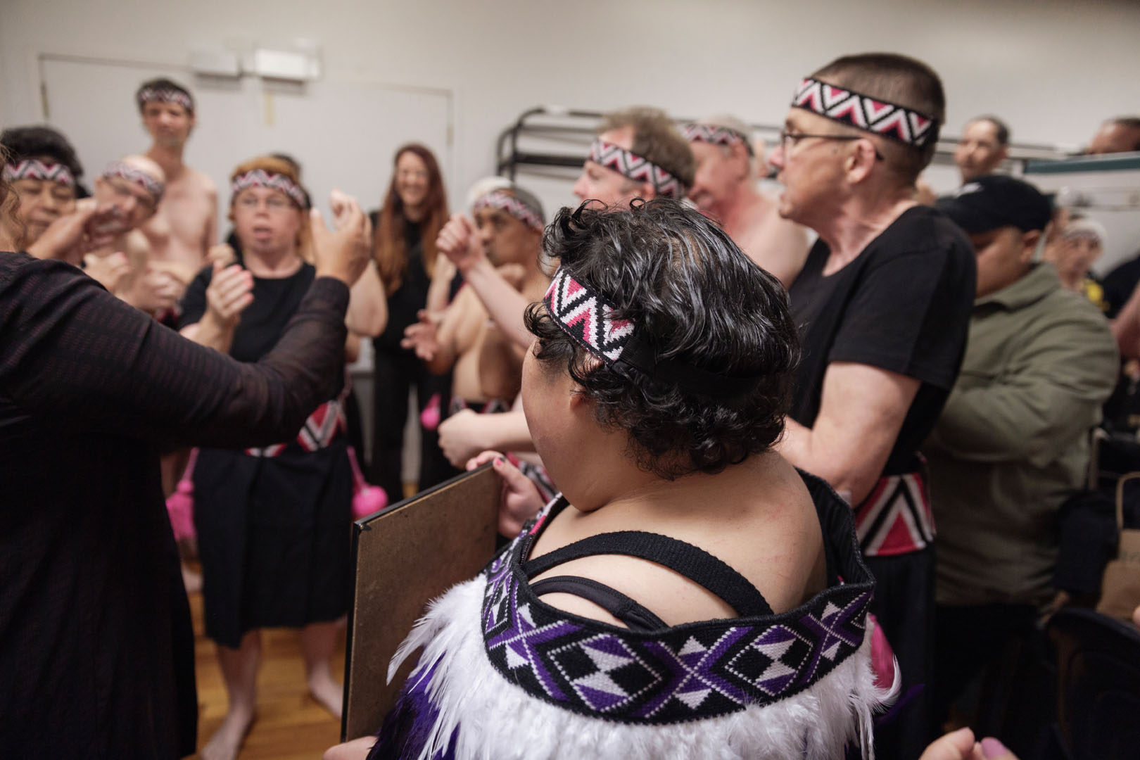 A group of kaihaka are practicing their moves backstage. The performers are wearing various garments including tīpare and a feather korowai.