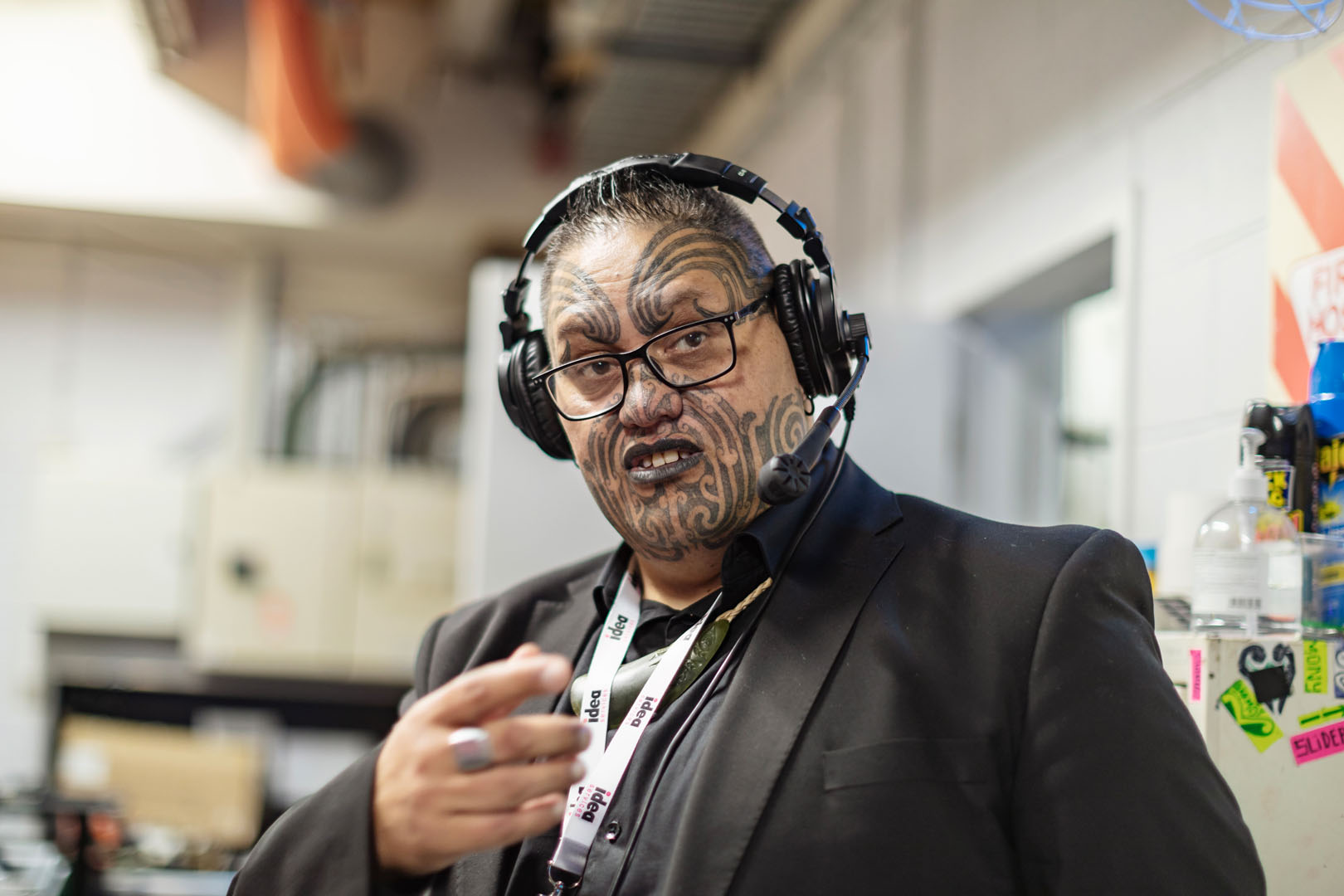 Taki Peeke wears a full face of ta moko, a headset with mic and a black suit. 