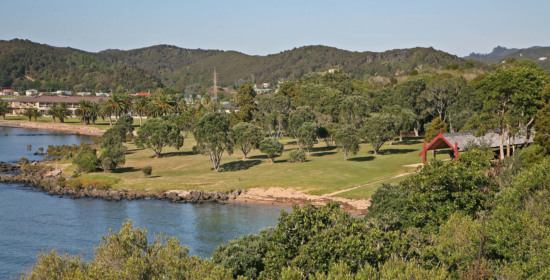 A green field with lots of trees by a body of water. A town and a Māori canoe is in the background