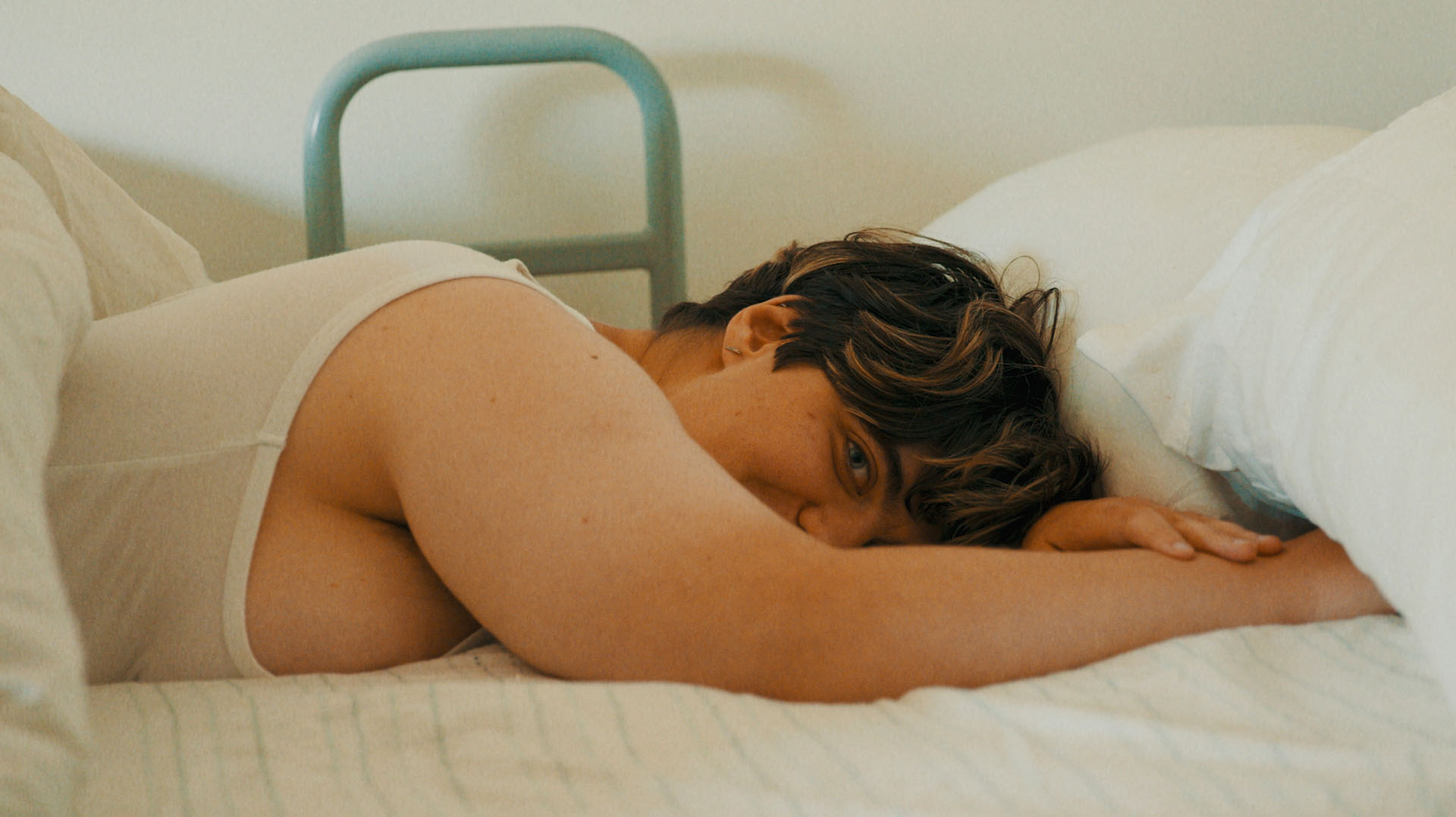 Henrietta Bollinger is a nonbinary person with short hair. They are lying in bed with white linen and smiles at the camera cheekily.