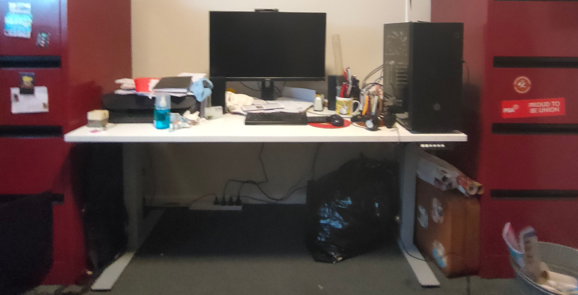 A white desk with a black monitor on top with various desk accessories, such as a keyboard, mouse, notepads and pens. Two red filing cabinets are on either side of the desk.