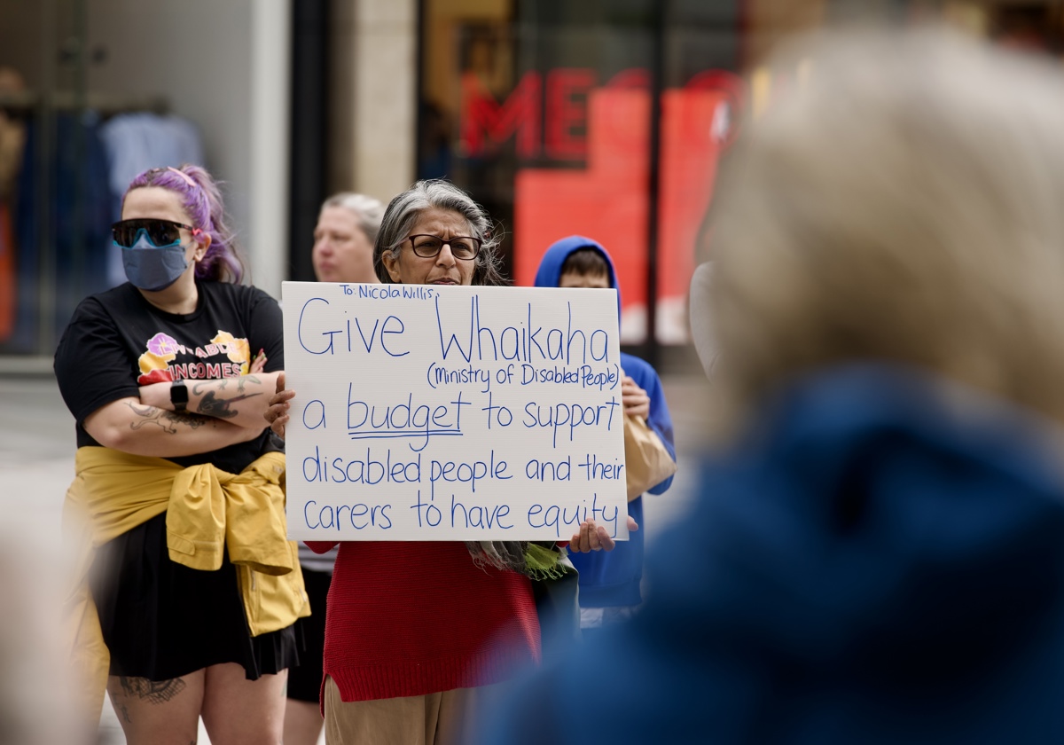 Anu Kialoti holds a white sign with dark blue writing which reads, “To: Nicola Willis, Give Whaikaha (Ministry of Disabled People) a budget to support disabled people and their carers to have equity”. 