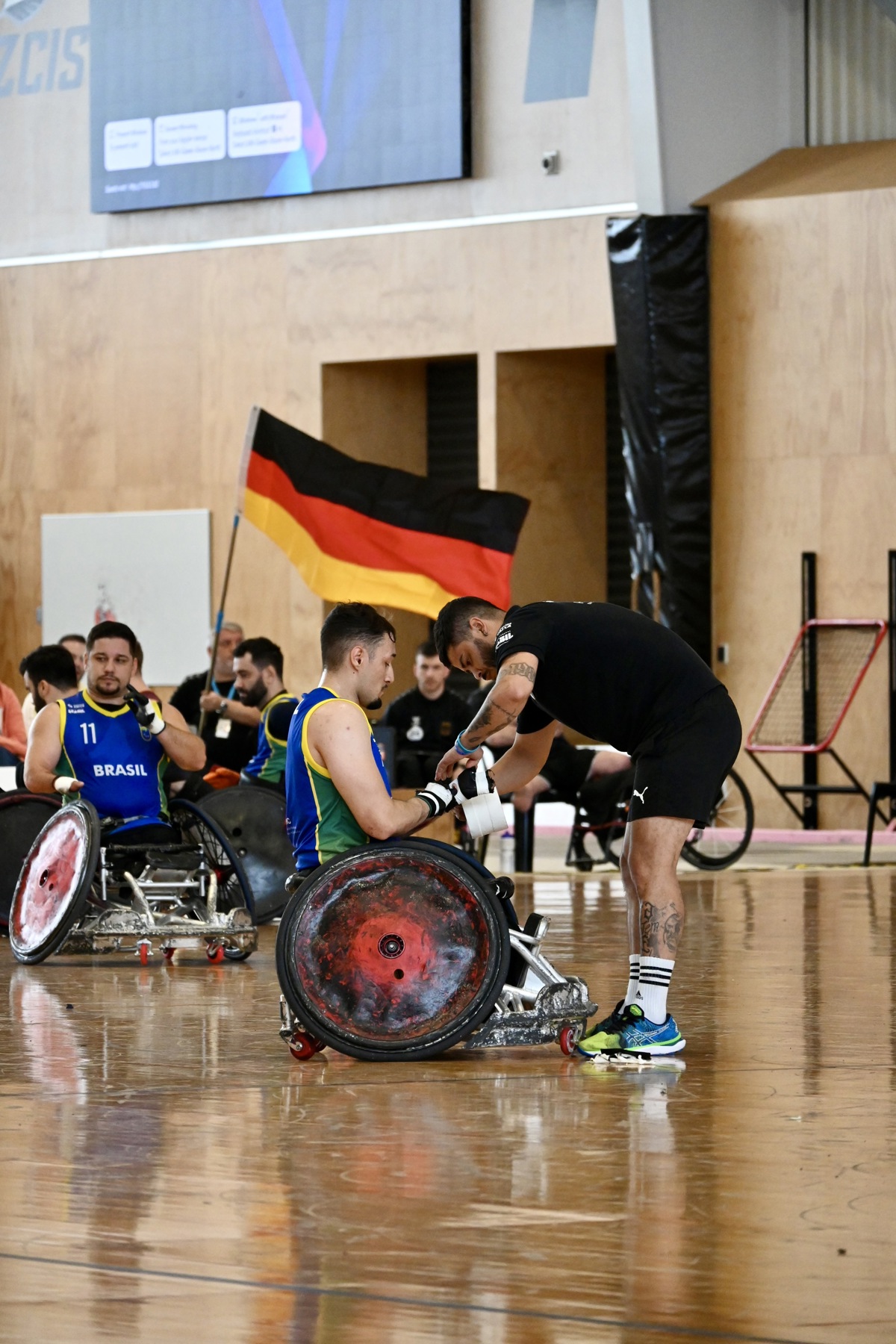 A Brazilian wheelchair rugby athlete has his hands wrapped up on the court. There is a Germany flag being waved behind him.