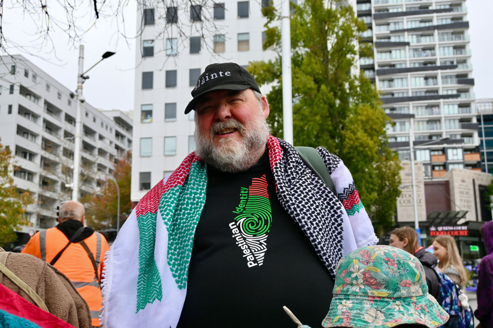 A man with a black cap and pro-Palestine clothing is at a protest in front of city buildings.