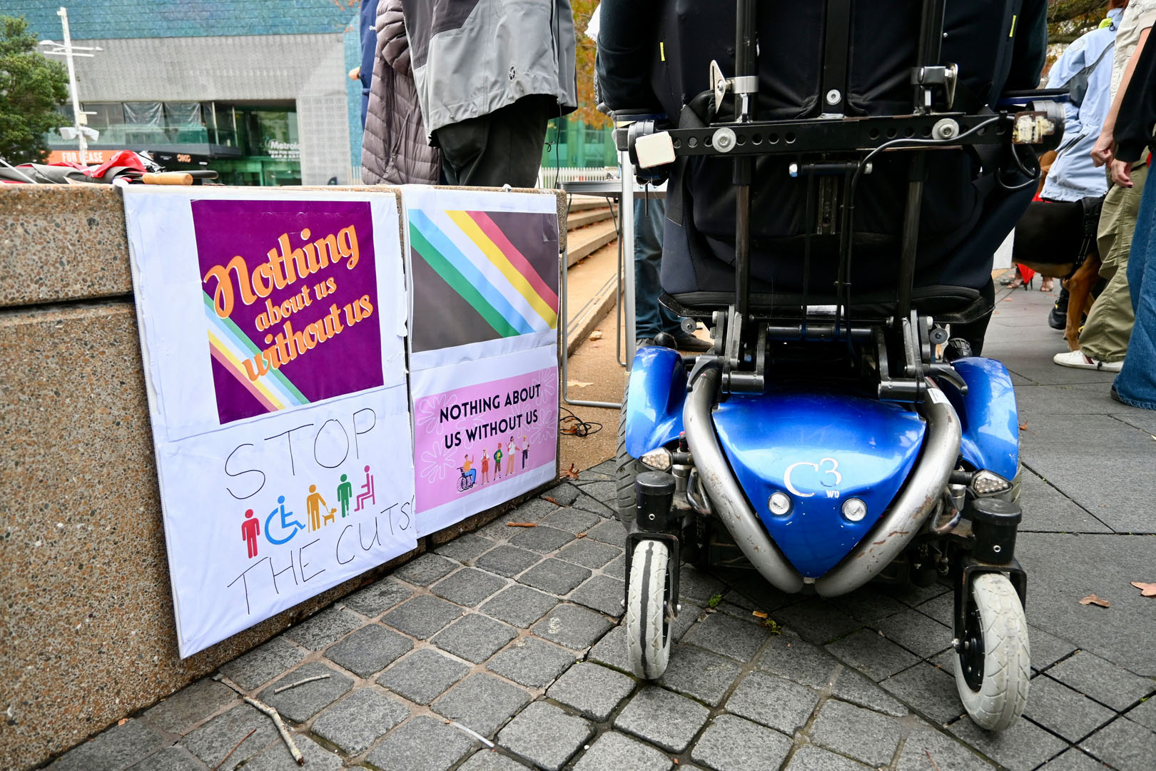 The wheels of a powered-wheelchair are rolling next to signs that read: Nothing about us without us, and Stop the cuts.