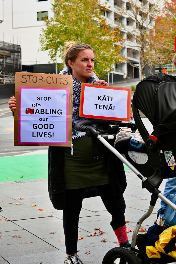 A woman with a prosthetic leg stands behind a baby pram and holds up two signs that read: Stop cuts, stop disabling our good lives! And: Kāti tēnā!