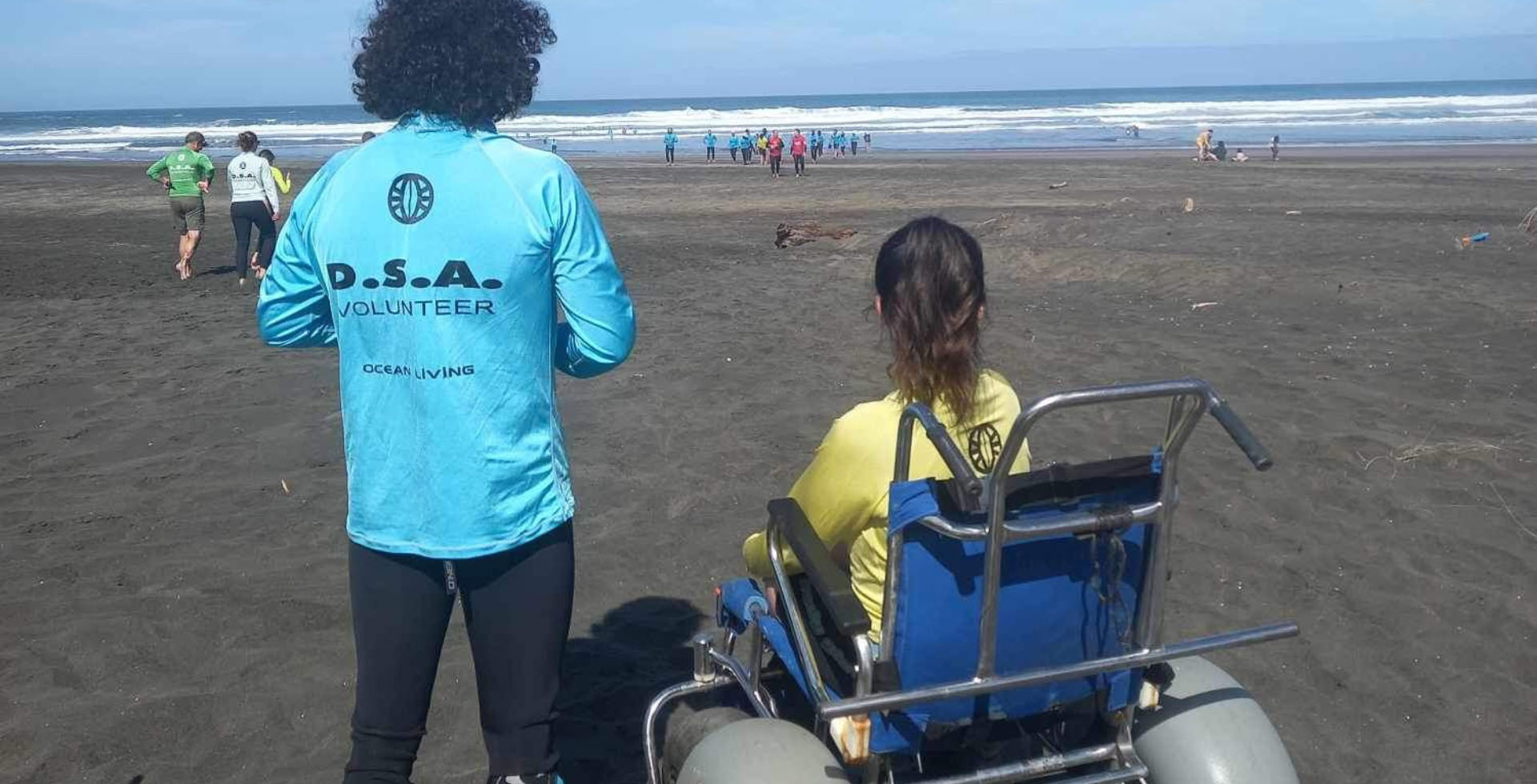 Olivia is sat in a big, beach wheelchair at Piha Beach; a volunteer stands next to her wearing a 'DSA Volunteer' rash shirt. They are both looking out at the waves.