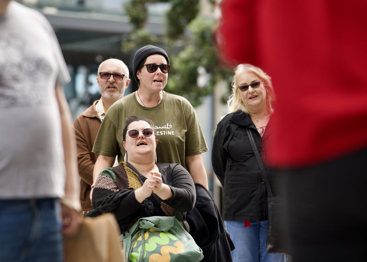 Image description: Danni, seated, is clapping while chanting with the crowd. Elle, who wears a khaki Palestine shirt, stands close behind Danni and chants along. 