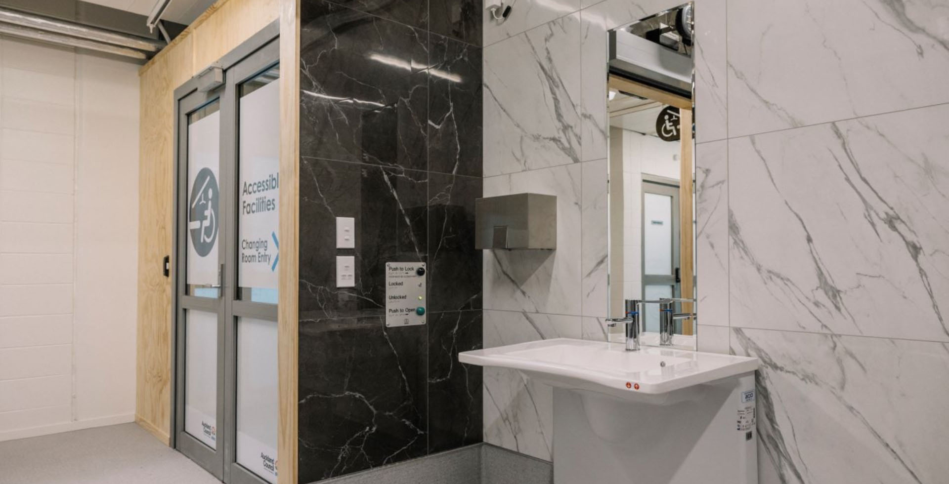 A corner of the spacious accessible changing room facility at Albany Stadium Pool shows sleek marble tiled walls. On the black marble side is a control panel for the automatic doors, and on the white marble side is a roll under sink under a long mirror. 
