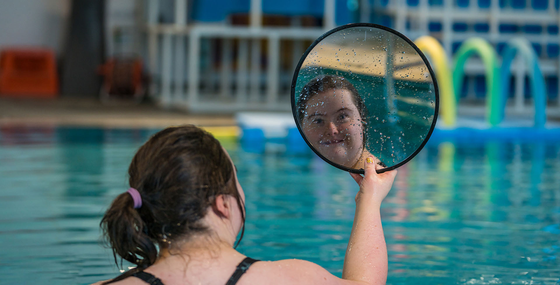 In the reflection of a circular hand-held mirror, a woman smiles while they enjoy a swim. A structure made of pool noodles floats in the background. 