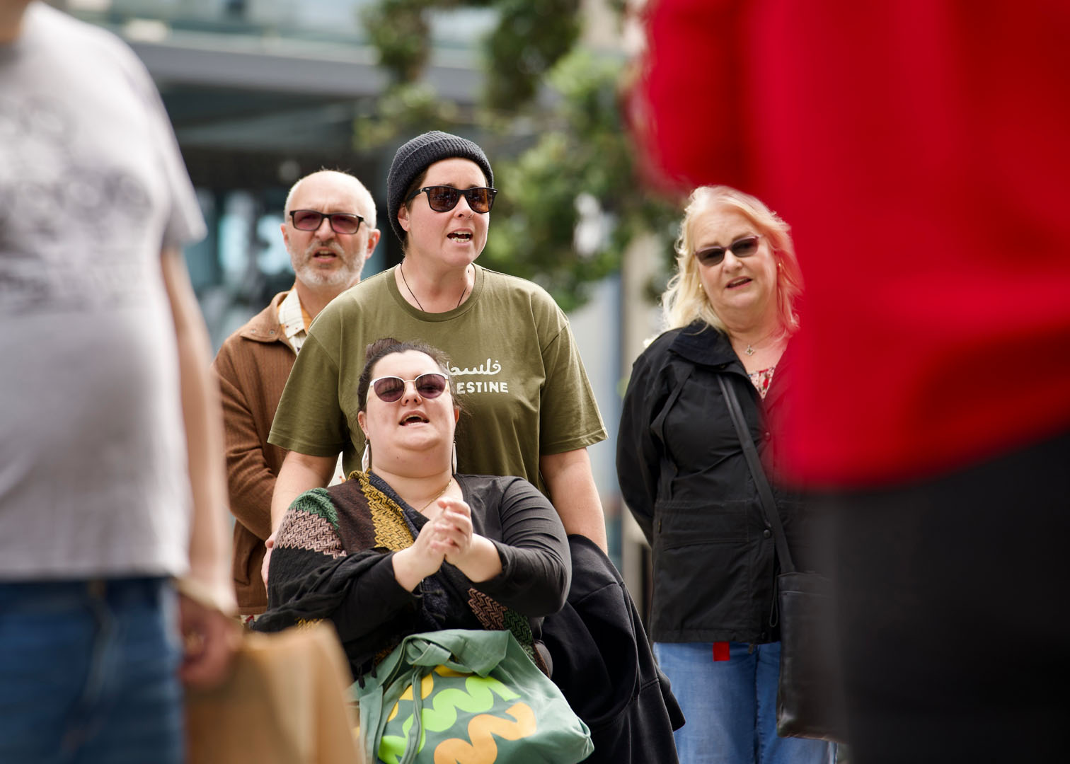 Image description: Danni, seated, is clapping while chanting with the crowd. Elle, who wears a khaki Palestine shirt, stands close behind Danni and chants along. 