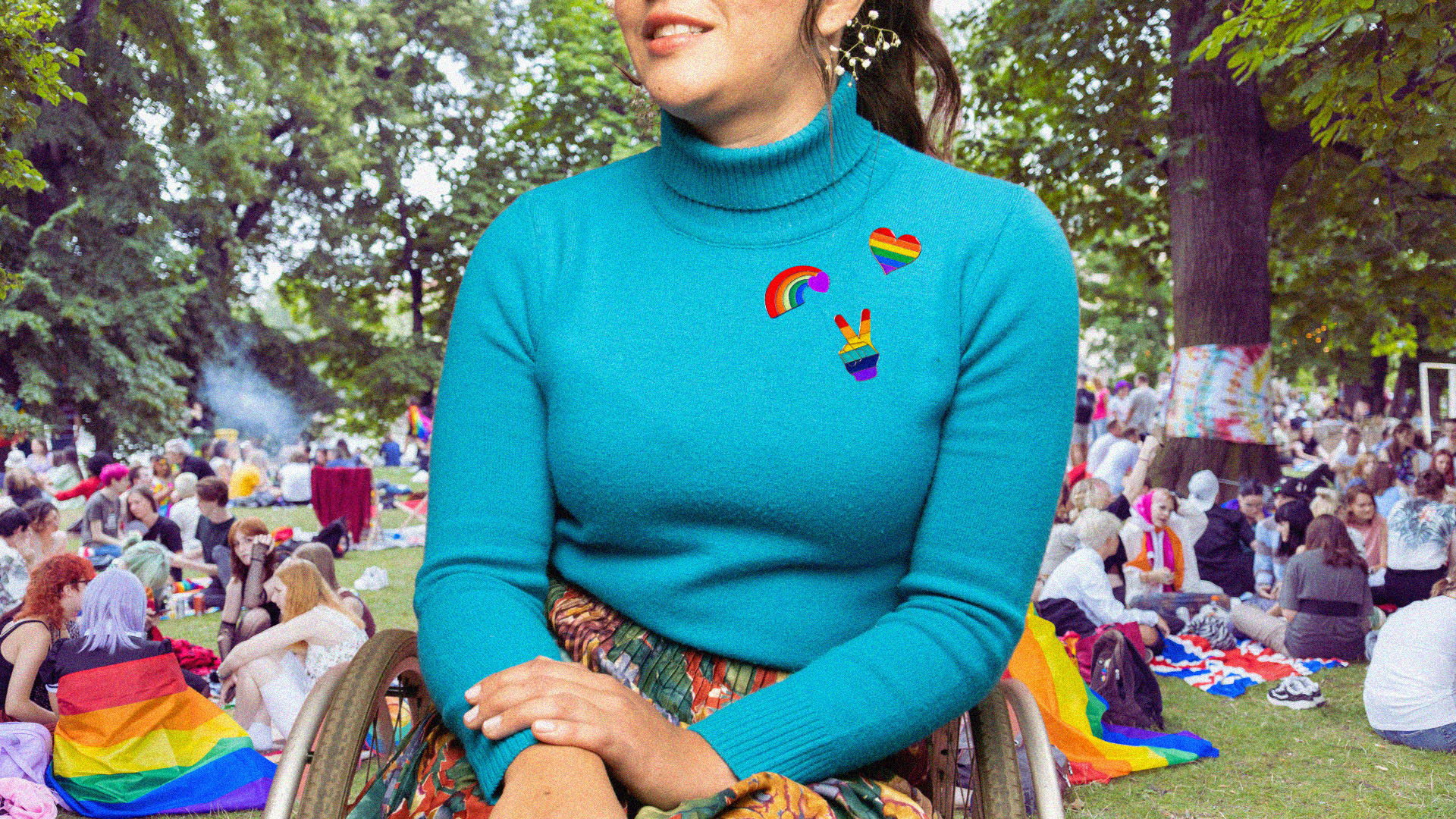 Person in wheelchair at a pride festival, they have pride stickers on their shirt in front of a crowd of festival goers.