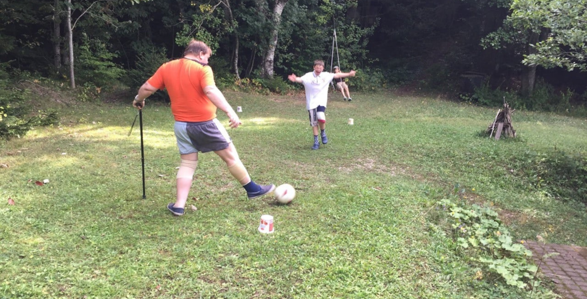 Robert wears a red shirt and shorts, while kicking a football with some young people on the grass.