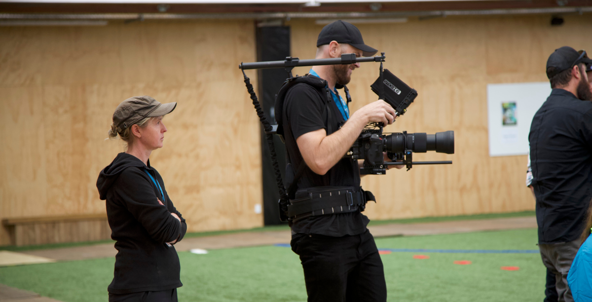 Robyn stands behind a camera operator in a sports gym.