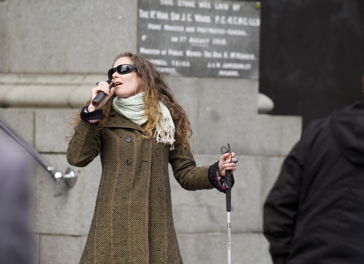 Caitlin Smith leans into a note as she sings into the microphone. Caitlin is wearing black sunglasses, a mint green scarf and a tweed coat. She holds a white cane in her left hand.