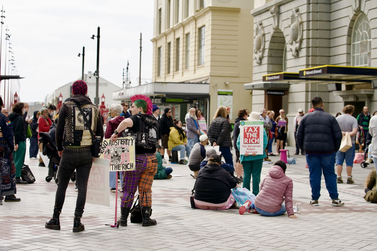 Protestors gather in the rally outside Britomart train station holding placards and watching speakers. Some are sitting on the floor of the paved square. 