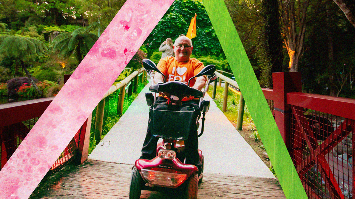 A person rides a mobility scooter across a bridge with wooden rails. There are strips of colour surrounding him.