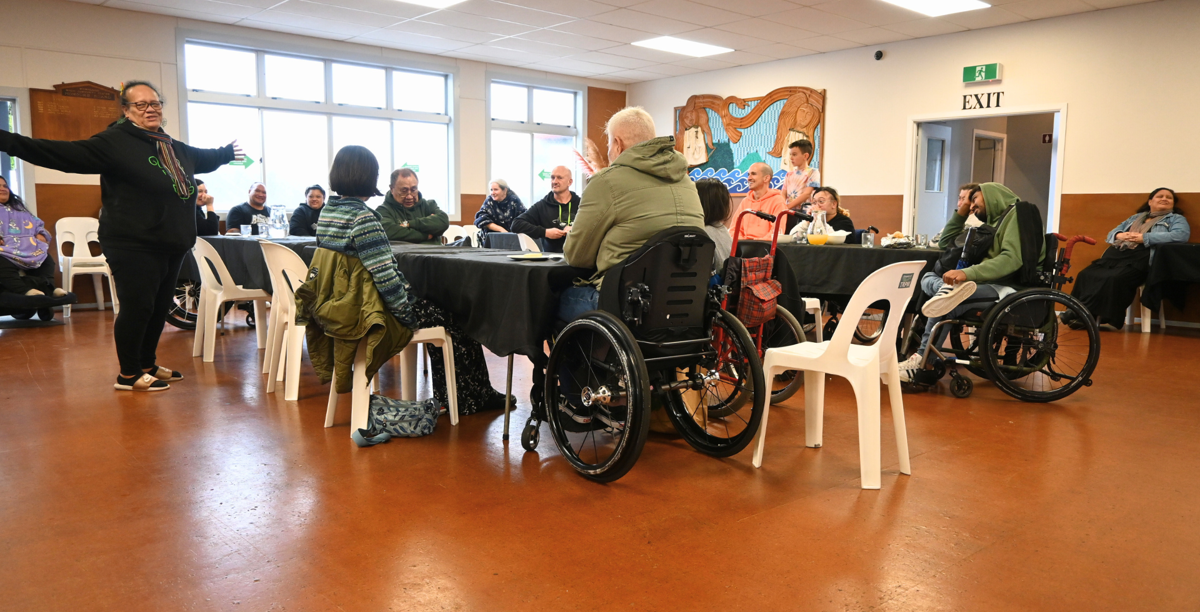 A group of people gather in a dining room, smiling, while listening to someone speak.