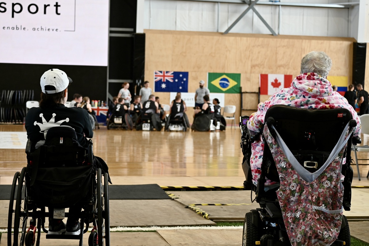Two people in wheelchairs are watching wheelchair rugby from the court side.