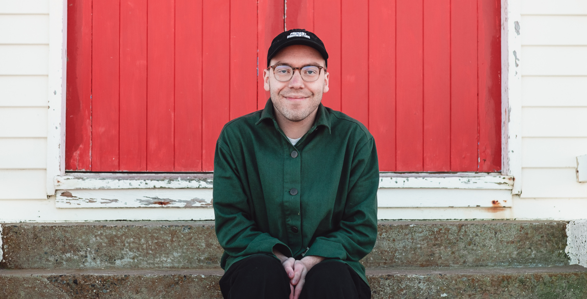 Jesse Austin-Stewart wears glasses, a black cap and a green shirt. He sits in front of a red door, smiling at the camera.