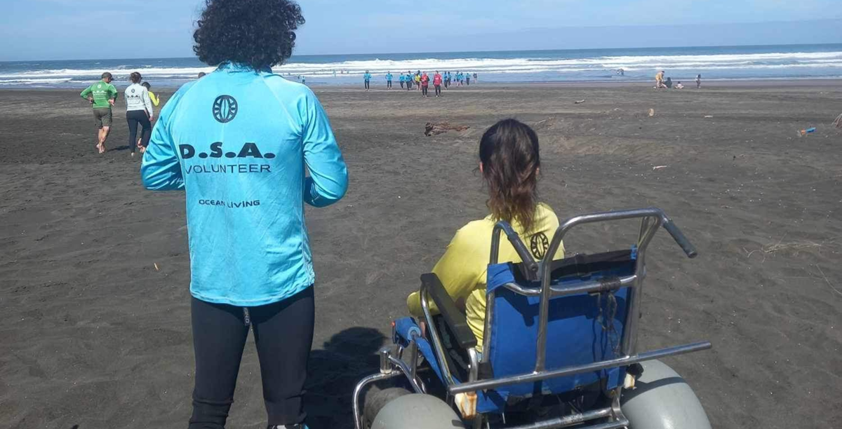 Olivia is sat in a big, beach wheelchair at Piha Beach; a volunteer stands next to her wearing a 'DSA Volunteer' rash shirt. They are both looking out at the waves.