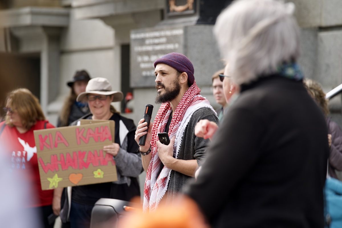 AJ, who is wearing a red and white keffiyeh and a purple beanie, speaks into the microphone. 