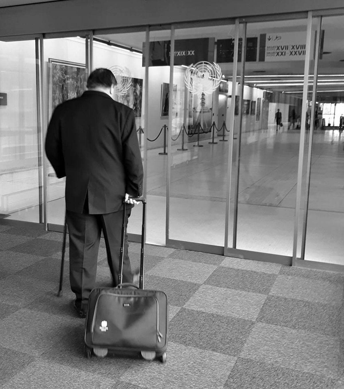 A black and white photo of Robert's back as she walks through some glass doors at the UN. He uses a walking stick and pulls along a small suitcase.
