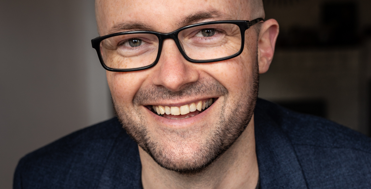 A close headshot of Steve smiling at the camera. He wears black frame glasses and a denim blue blazer. 