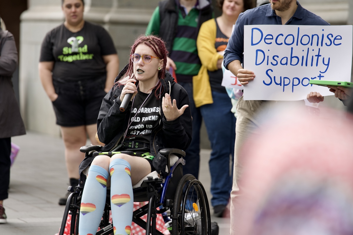 Keiko, who uses a wheelchair, addresses the rally. They have burgundy dreadlocks and are wearing knee high blue socks with rainbow hearts on them.