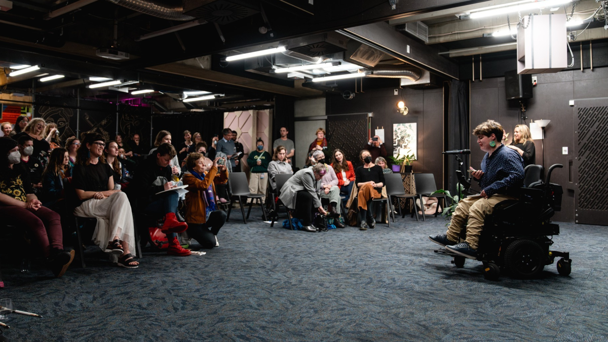 Image description: An audience of disabled and non-disabled people are mesmerised by Henrietta reading from their book. 
