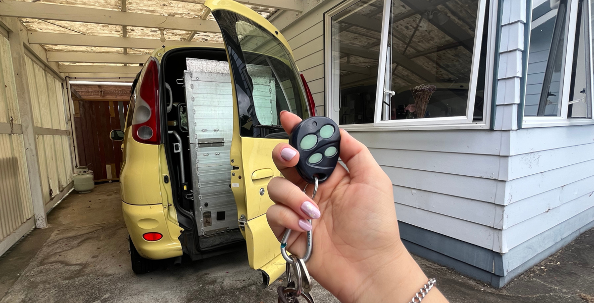 A hand holds up a set of car keys with a small remote control. In the background is a yellow car with the backdoor ajar and a metal wheelchair ramp.