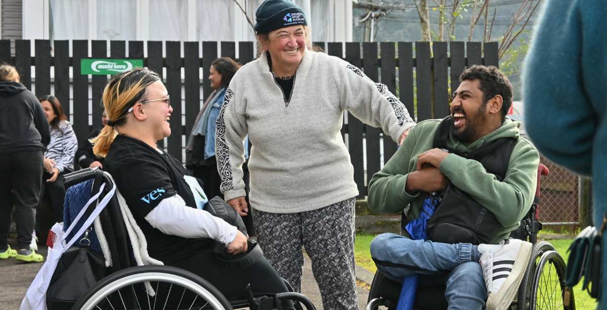 Two young wheelchair-users and a person standing smile and laugh while chatting to each other.