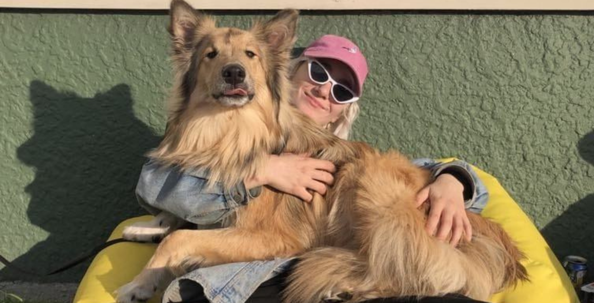 Asia reclines on a yellow bean bag outside in front of a green textured wall on a gingham red picnic mat. She cuddles a big dog as she smiles at the camera. 