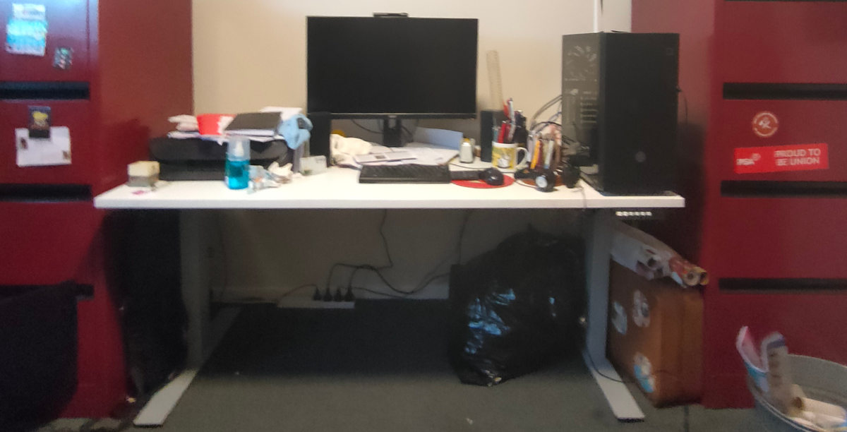 A white desk with a black monitor on top with various desk accessories, such as a keyboard, mouse, notepads and pens. Two red filing cabinets are on either side of the desk.