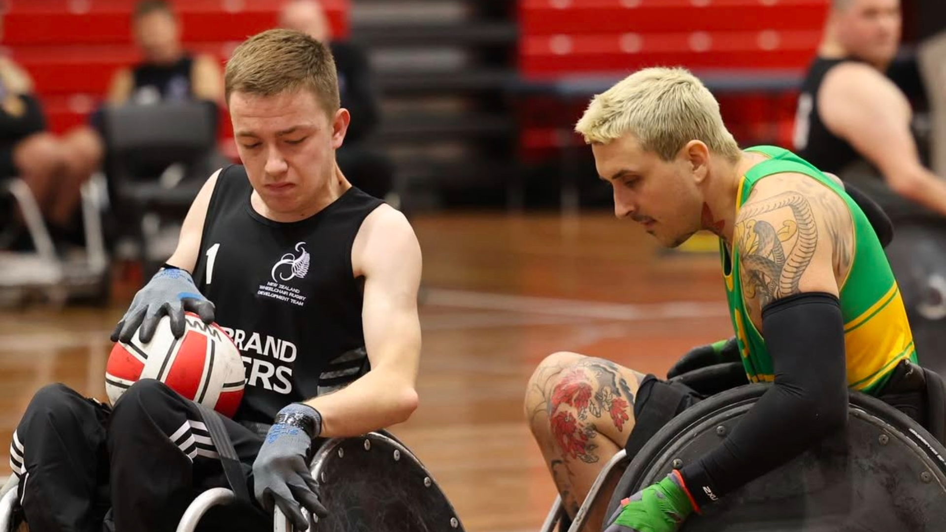 Dylan, wearing a black New Zealand jersey, plays wheelchair rugby on a court holding a ball. He is near an opponent in a bright yellow and green jersey, both engaged in the game