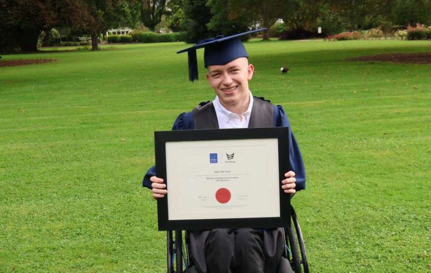 Dylan smiles on a green field in a royal blue graduation gown, proudly holding his degree.