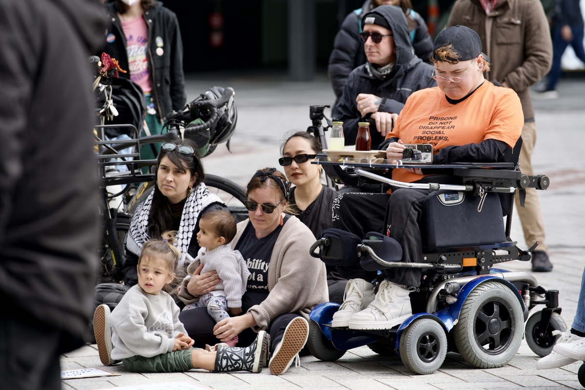 Robbie and her two young children sit on the pavement surrounded by other protestors. Camille, who uses a power chair, films the speeches next to her. 