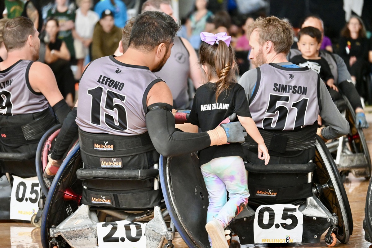 A small child stands between two wheelchair athletes, with a player wearing a singlet reading "LEFONO 11" has his hand wrapped around her.