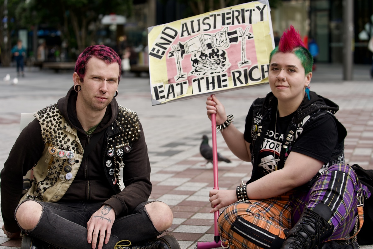 A pair with bright coloured hair sitting on the floor of the square holding a sign propped on the end of a pink walking stick with the monopoly man on a spit roast between “END AUSTERITY EAT THE RICH”. 