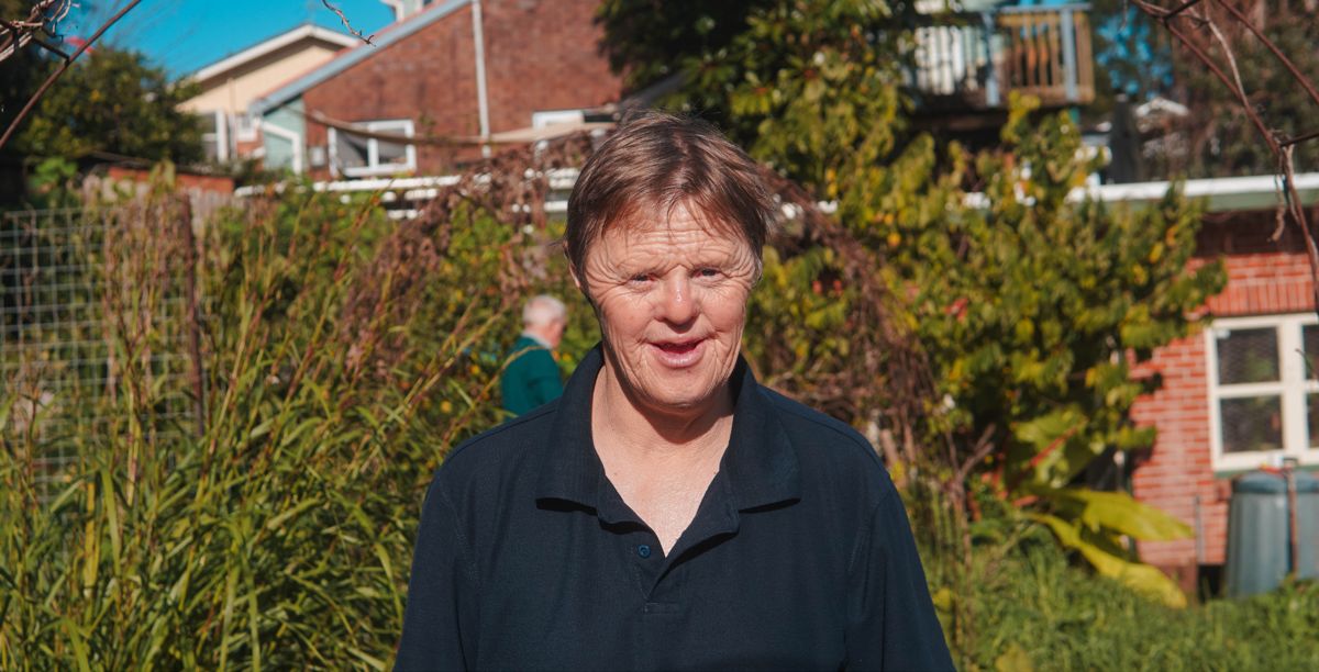 Garden volunteer Daniel Clarke wears a black shirt and smiles at the camera; in the background is a garden with green bushes and trees.