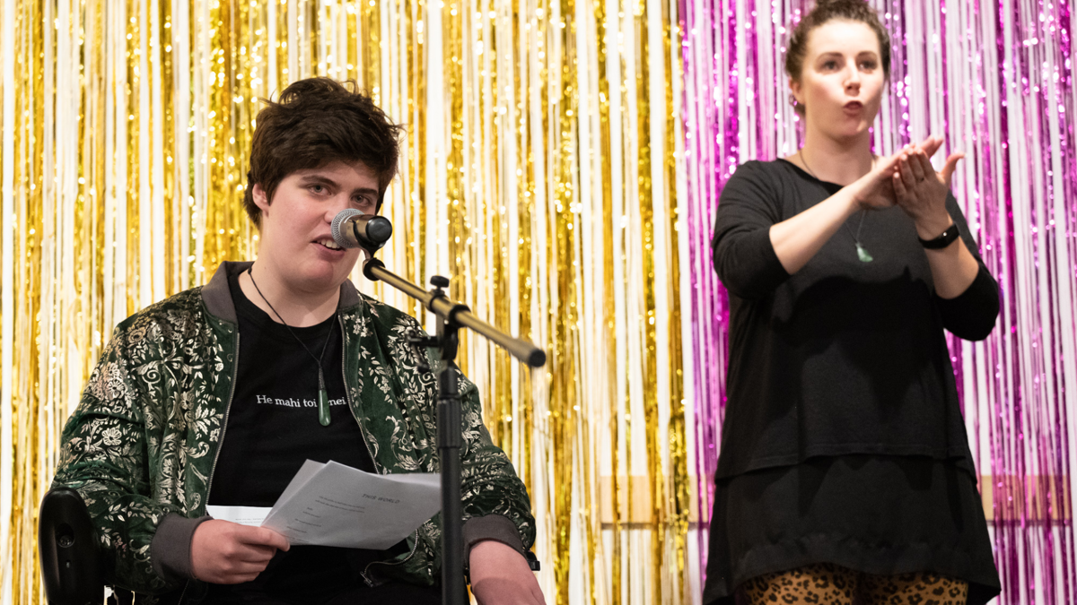 Henrietta is a pākehā person with short brown hair; they are speaking into a microphone. A sign language interpreter is beside them,